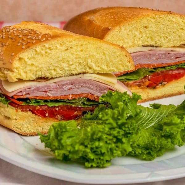 Two halves of a deli sandwich with ham, cheese, lettuce, and tomatoes, served with a side of lettuce.