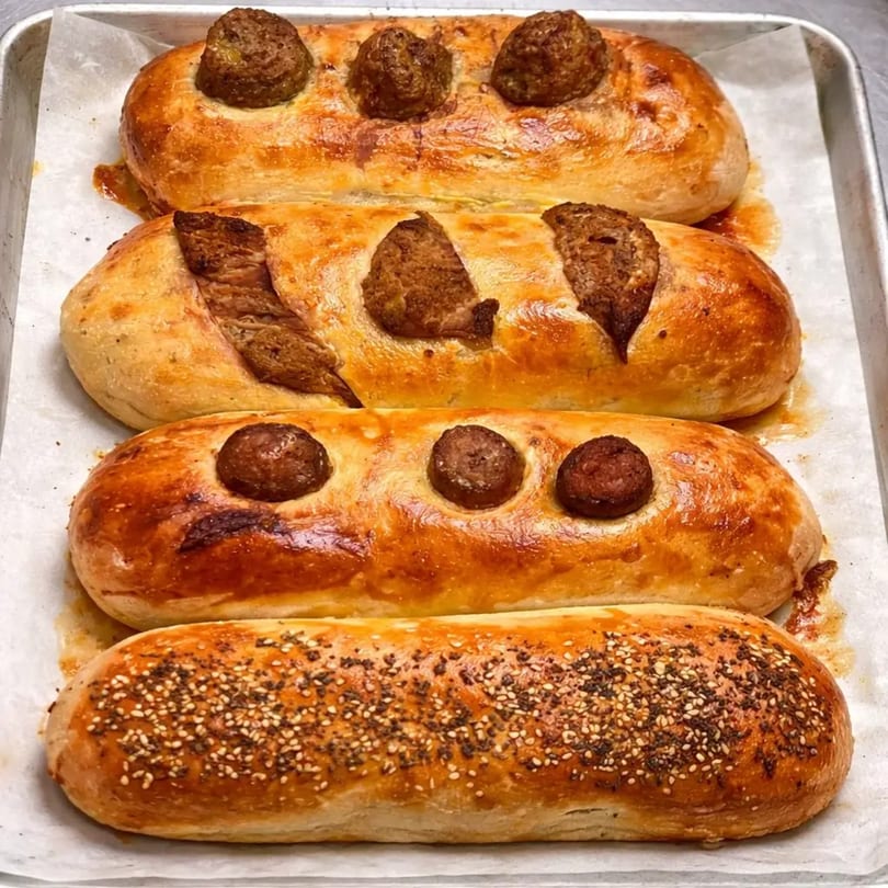 Four golden-brown baked bread loaves, some with meatballs and one with sesame seeds, on a baking sheet.