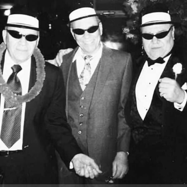 Three men in hats and sunglasses, two in suits and one in a tuxedo, posing together.