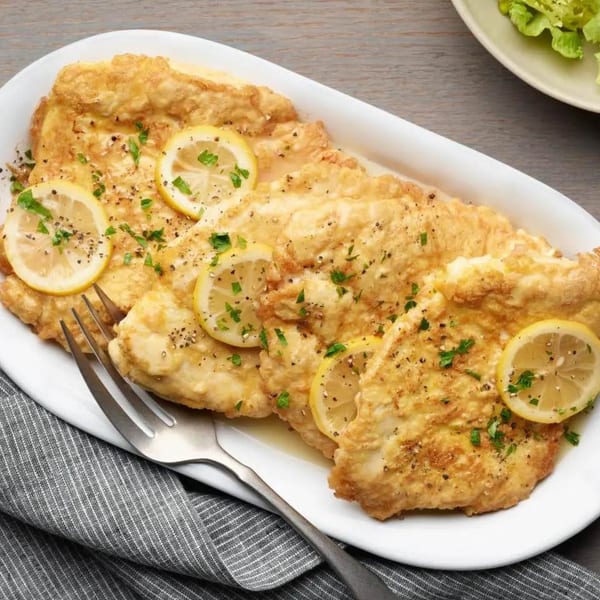 Golden-brown chicken cutlets with lemon slices, parsley, and black pepper on a white platter.