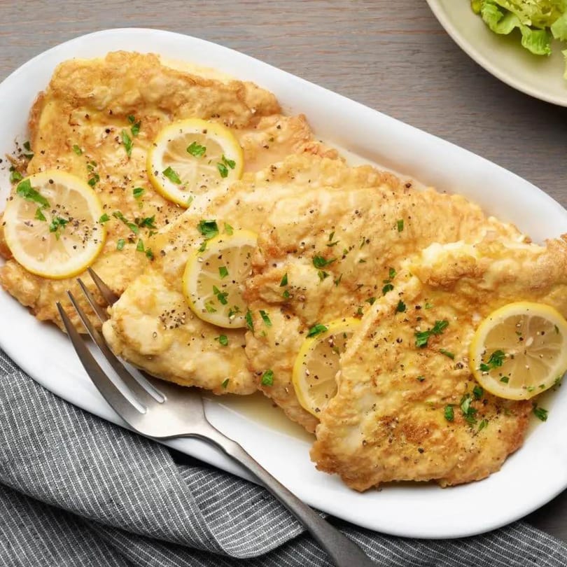 Golden-brown chicken cutlets with lemon slices, parsley, and black pepper on a white platter.